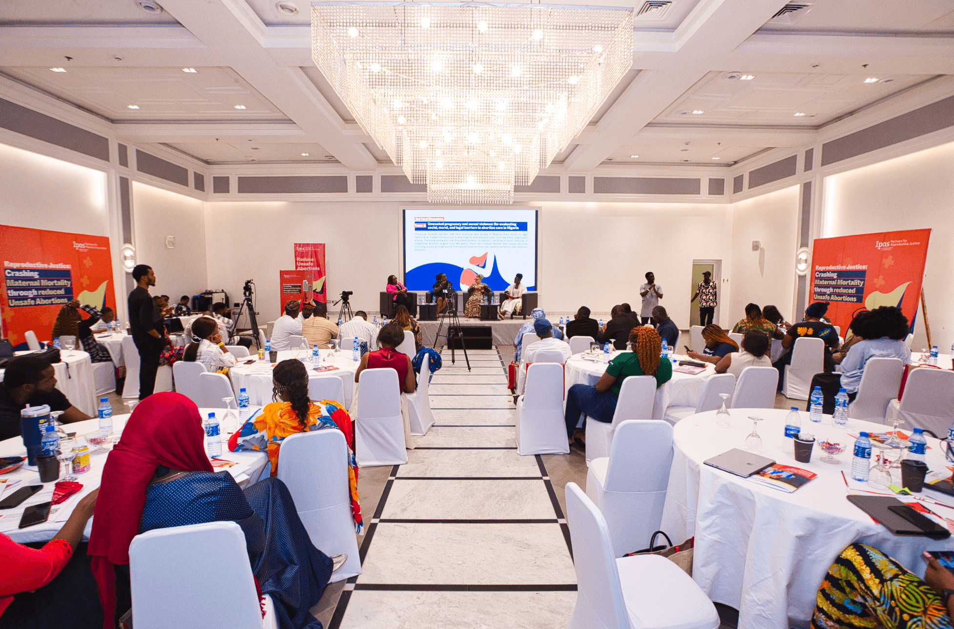 Wide view of a formal gala in a bright ballroom with a panel on stage and guests seated at round tables